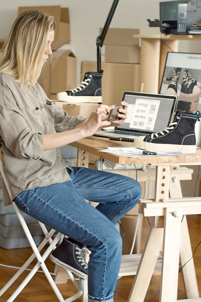 Woman using laptop and smartphone to manage an online shoe store in a workspace filled with boxes.