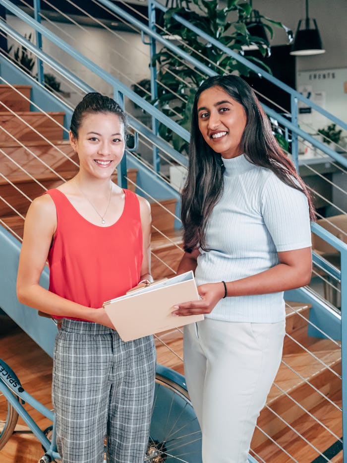 creative-03 Two young businesswomen collaborating in modern office space, standing by stairs, holding documents.