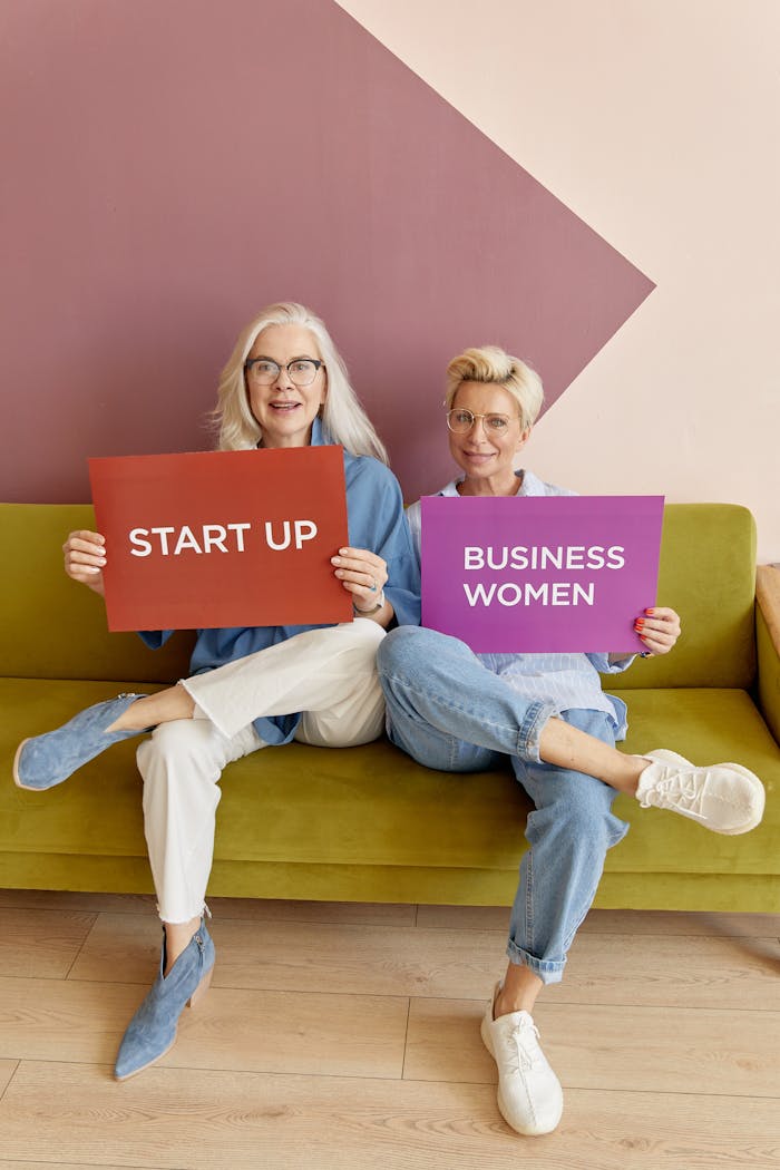 creative-02 Two senior businesswomen sitting on a couch, holding start-up placards with confidence.